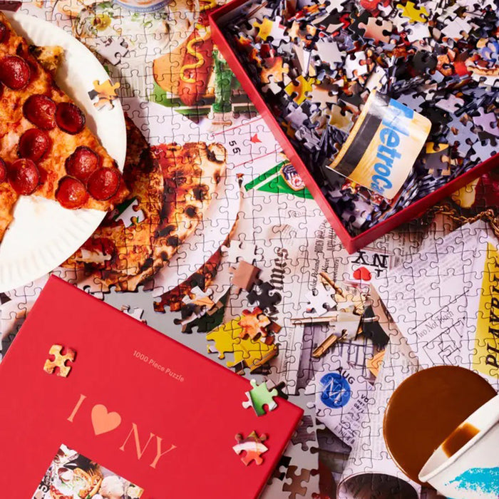 Piecework puzzles I heart NY 1000 piece NYC themed jigsaw puzzle in red box, front view with partially completed puzzle in the background, a slice of pizza, a metrocard and a spilled coffee.