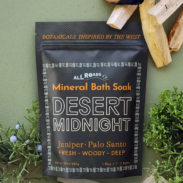 A black stand-up pouch of All Roads Mineral Bath Soak, flavor Desert Midnight, styled on a textured sage green background with palo santo sticks and juniper sprigs.