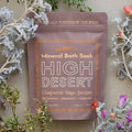 A stand-up dusty pink pouch of All Roads High Desert Mineral Bath Soak, styled with dried chaparral, sage, and juniper flowers against a neutral beige background.