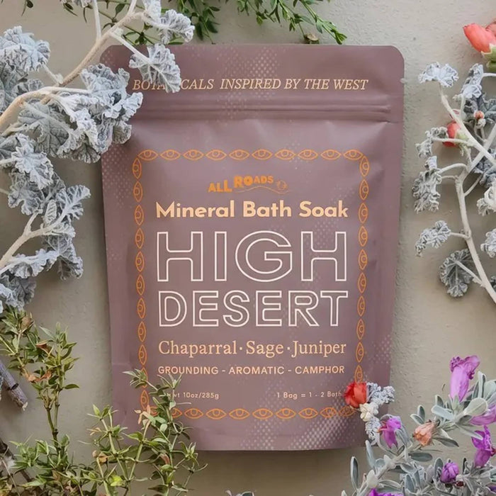 A stand-up dusty pink pouch of All Roads High Desert Mineral Bath Soak, styled with dried chaparral, sage, and juniper flowers against a neutral beige background.