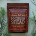 A deep burgundy stand-up pouch of All Roads Home Stead Mineral Bath Soak, surrounded by pine needles on a light sage green background.
