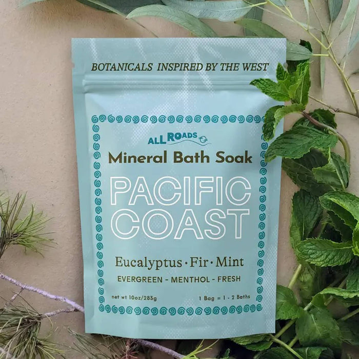 A stand-up pouch of All Roads Pacific Coast Mineral Bath Soak with Eucalyptus, Fir, and Mint scent. The pouch is a pale blue with teal and deep green text, styled on a beige textured background with green eucalyptus leaves and pine needles.