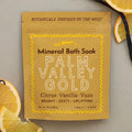 A yellow pouch of All Roads Palm Valley Gold Mineral Bath Soak, displayed on a textured gray background with decorative citrus slices and a vanilla bean.