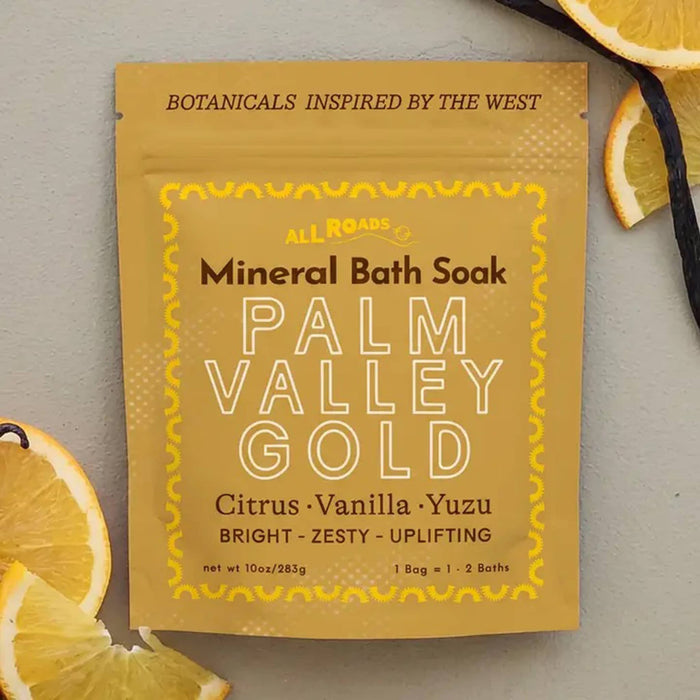 A yellow pouch of All Roads Palm Valley Gold Mineral Bath Soak, displayed on a textured gray background with decorative citrus slices and a vanilla bean.