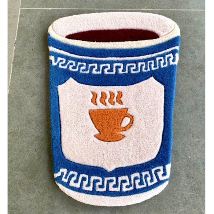 A tufted accent rug shaped like a cup with the Anthora coffee cup design, including a blue and white Greek key pattern and a central shield design featuring an orange coffee cup with steam, shown on a gray tile floor.