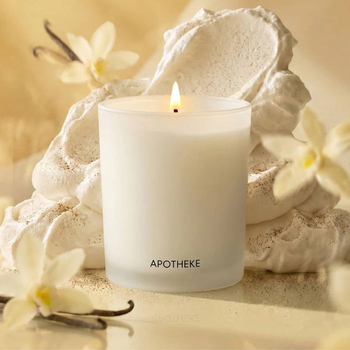White lit candle with 'Apotheke' branding on a yellow background with vanilla beans, flowers and whipped cream.