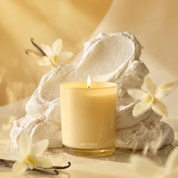 Lit candle with 'Apotheke' branding on a yellow surface with flowers, vanilla beans and whipped cream.