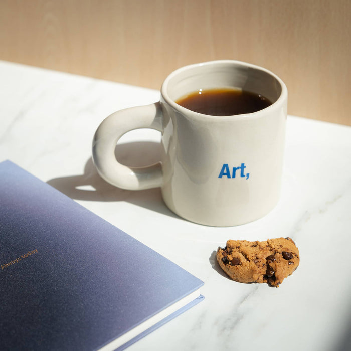 White mug with 'Art,' text, a cookie, and a notebook on a light surface.