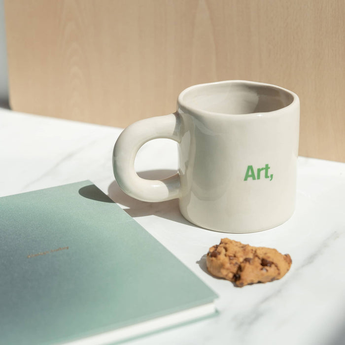 White mug with 'Art,' text, a cookie, and a notebook on a light surface.