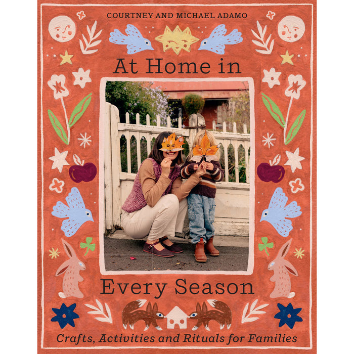 The front cover of the book 'At Home in Every Season' by Courtney and Michael Adamo. The cover features a terracotta orange background decorated with whimsical folk-art illustrations of bunnies, birds, flowers, and stars, surrounding a central color photograph of a woman and child wearing orange paper hats.