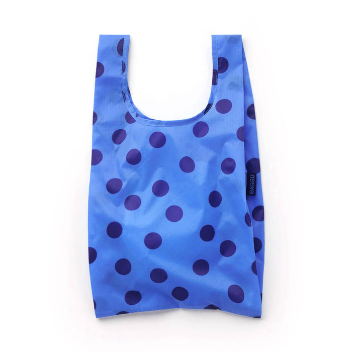 A reusable folding shopping bag in bright blue with dark blue polka dots, shown against a plain white background. A small black tag on the side displays the brand name 'BAGGU'.