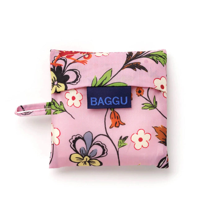 A pink reusable bag with a colorful floral print  in matching pouch with a navy blue brand label reading 'BAGGU' on the front flap, photographed on a clean white background.