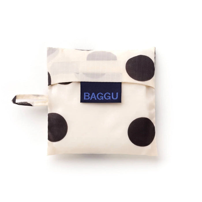 A folded reusable bag from BAGGU, in a cream-colored pouch with large black polka dots and a navy blue brand label on the front.