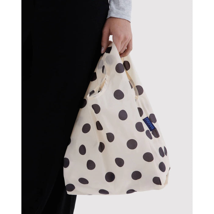 A person holding a reusable ripstop nylon tote bag with a cream base and black polka dots. The person is wearing a black skirt and a light gray long-sleeve top.
