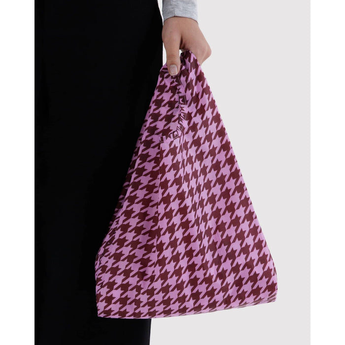 A person holding a tote bag with a pink and brown houndstooth pattern against a light gray background.