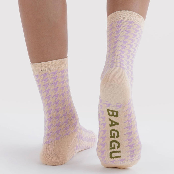 A pair of crew socks with a lilac and beige houndstooth pattern, worn on a model's feet against a light gray background. The brand name 'BAGGU' is printed vertically in olive green on the sole of the right sock.