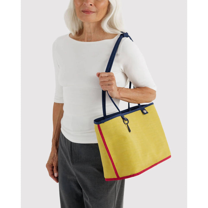 A woman wearing a white top and gray pants carries a yellow tote bag with navy blue straps and red trim against a light gray background.