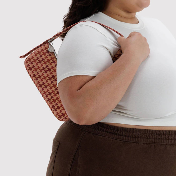 A person wearing a white t-shirt and brown pants carries a patterned tan and red shoulder bag over their shoulder against a light gray background.