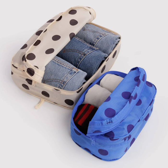 2 recycled nylon zippered packing cubes, large bag is cream with black polka dots and jeans inside, smaller bag is blue with navy polka dots and socks inside, on a white background.
