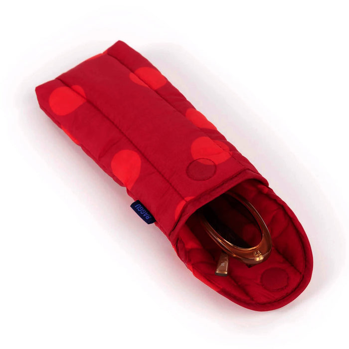 A red quilted fabric glasses pouch with a large polka dot pattern in shades of red, featuring a fold-over flap and a small dark blue brand tag on the side, lying down and open with sunglasses inside on a white background.