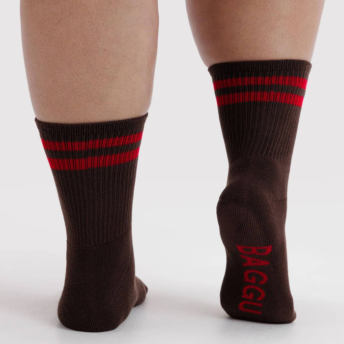 A pair of dark brown crew socks with red stripes near the cuff and red branding on the sole, shown on a model's feet against a light gray background.
