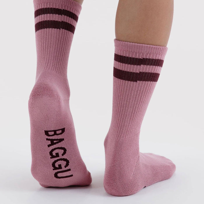 A pair of plum crew socks worn on legs, featuring two burgundy stripes near the cuff and the brand name 'BAGGU' printed in burgundy on the sole of one sock, against a plain light gray background.