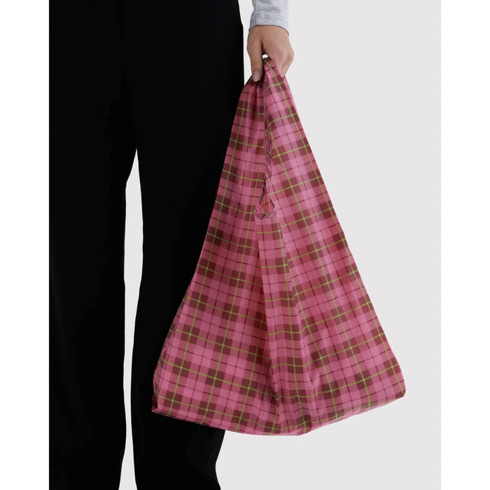 A person wearing black pants holds a reusable fabric shopping bag with a pink and lime green tartan pattern against a light gray background.