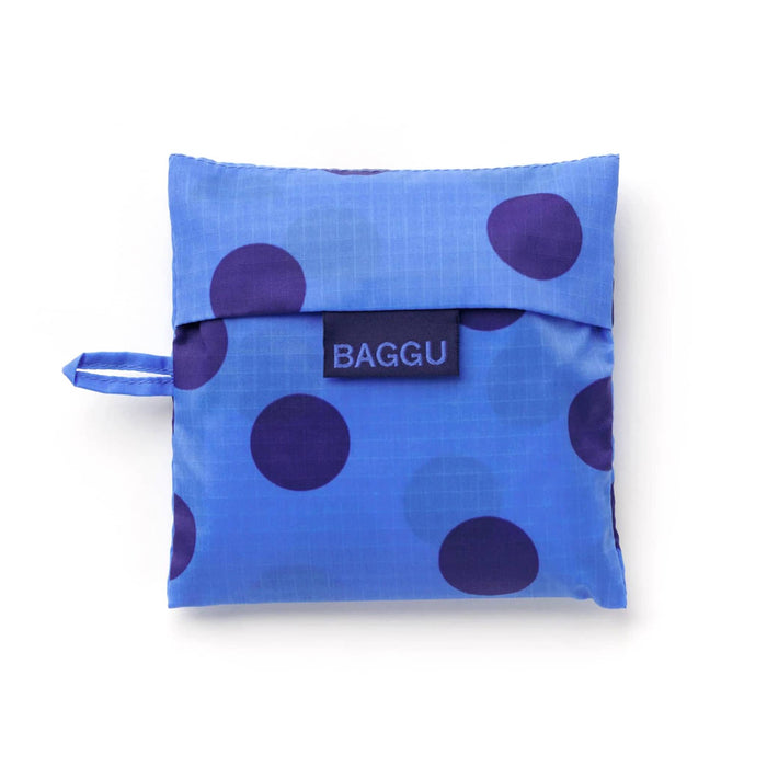 A folded reusable bag in a matching blue pouch with dark blue polka dots and a navy brand label reading 'BAGGU' on the front, shown on a clean white background.