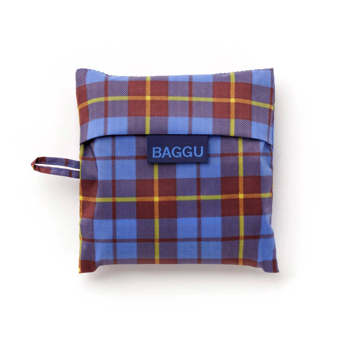 A folded reusable bag in a matching blue, red, and yellow tartan plaid pouch, featuring a navy blue flap with the brand name 'BAGGU' in white text and a matching plaid loop handle on the left.