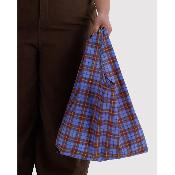 A person wearing dark brown pants holds a reusable fabric shopping bag with a blue, red, and yellow plaid pattern against a light gray background.