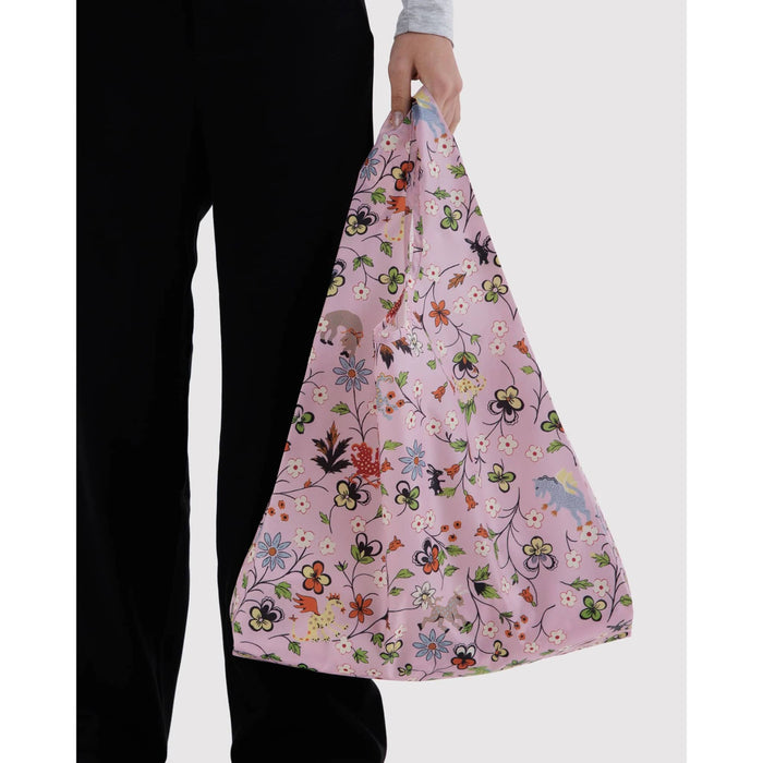 A person wearing black pants holds up a light pink reusable shopping bag with a colorful floral and mythical creatures pattern against a plain light gray background.