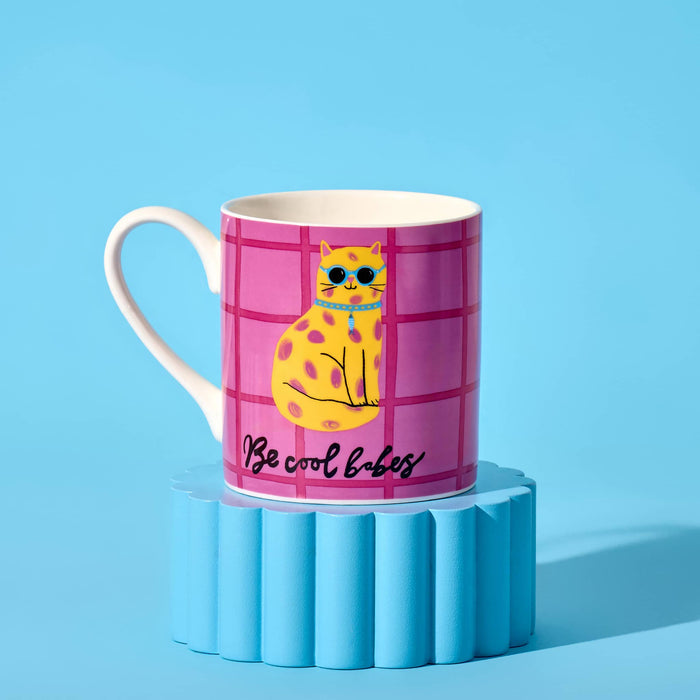 A white ceramic mug featuring a pink grid pattern with a yellow spotted cat wearing blue sunglasses and the text 'Be cool babes' in black script. Handle is on the left and it's sitting on a blue ribbed pedestal against a light blue background.
