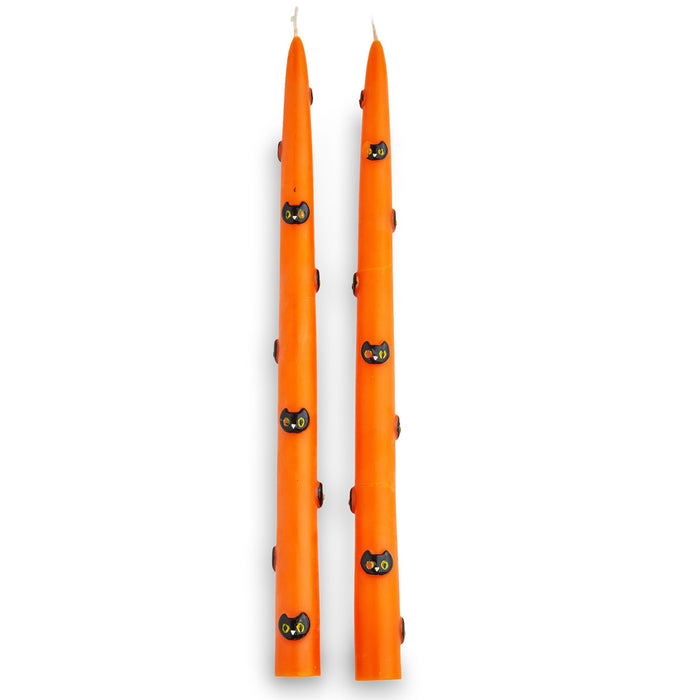 A pair of tall orange taper candles decorated with small black cat faces, shown on a plain white background.