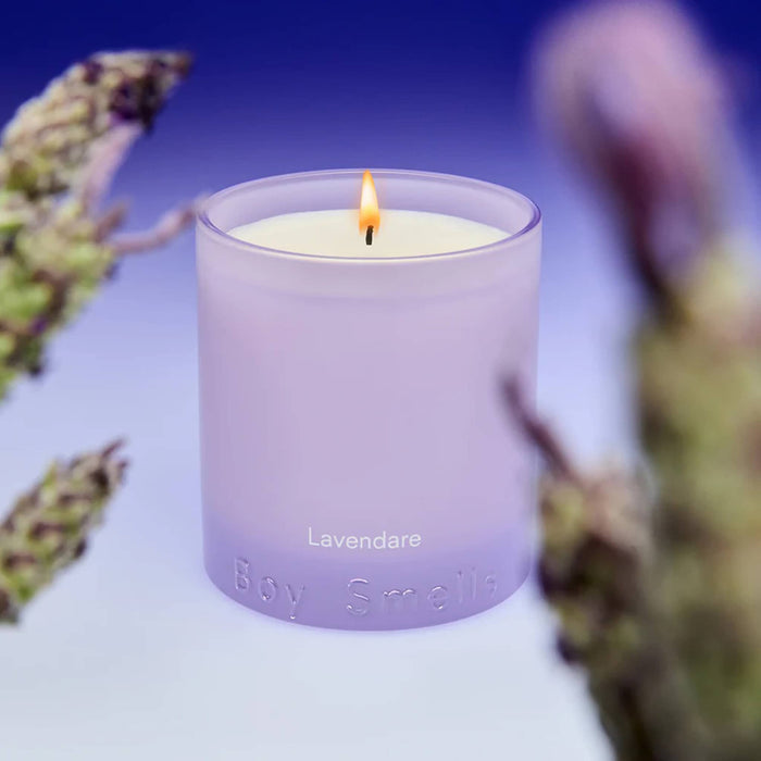 Boy Smells Lavendare scented 9 ounce Essentials candle in lavender vessel, lit, with lavender blooms out of focus in foreground.