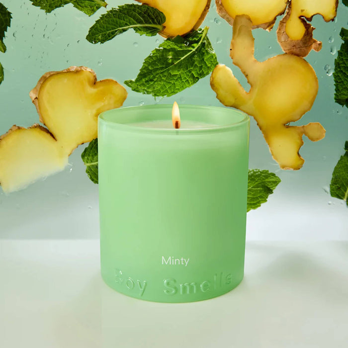 Boy Smells Minty scented 9 ounce Essentials candle in light green glass vessel, lit, with mint and ginger in the background.