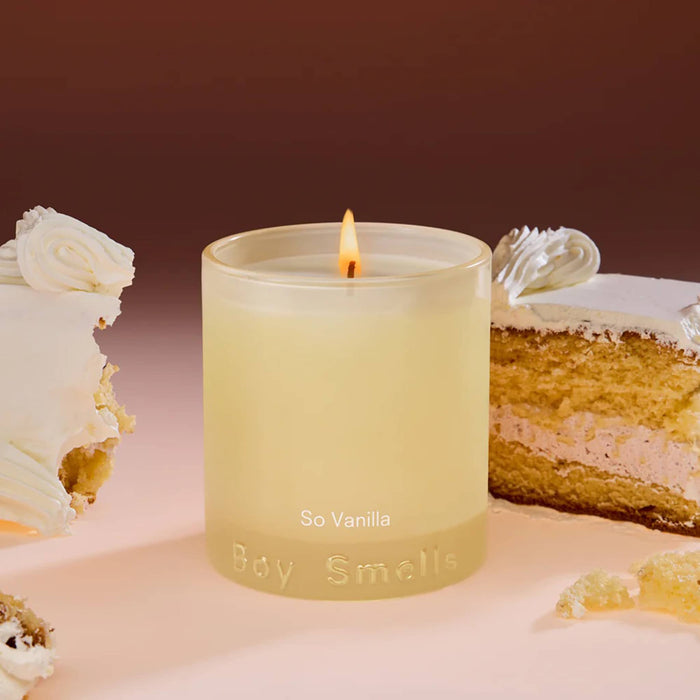 Boy Smells So Vanilla scented 9 ounce Essentials candle in cream glass vessel, lit, with slices of vanilla cake in the background.