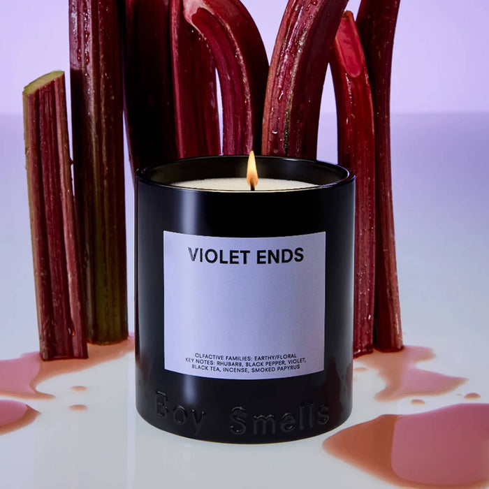 Boy Smells Violet Ends scented 9 ounce Classics candle in black glass vessel with dark violet label, lit with rhubarb stalks in the background.