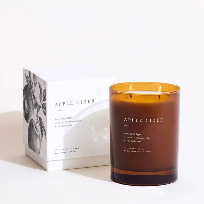An amber glass jar candle labeled APPLE CIDER, shown next to its white box packaging with a black botanical illustration, on a clean white background.