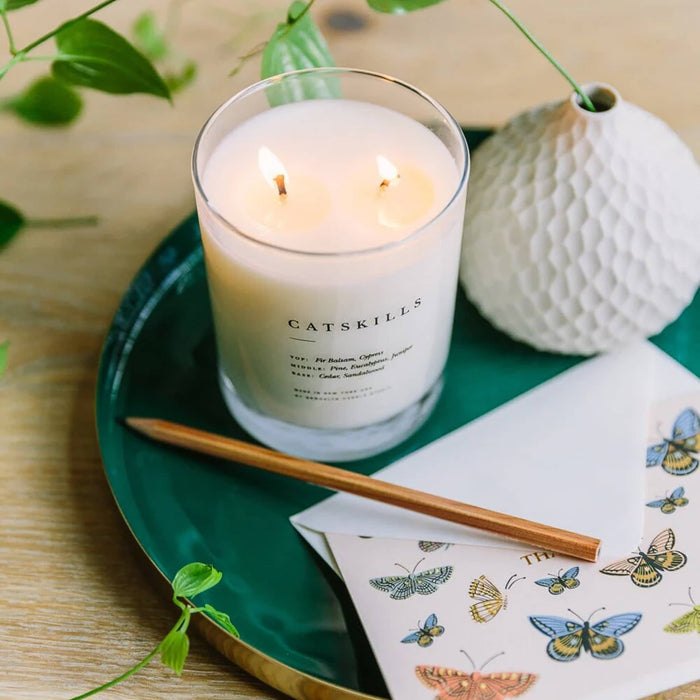 Brooklyn Candle Studio Catskills scented soy wax candle in clear glass vessel with double wicks, sitting on green tray with white vase, greeting card and wicks are lit.