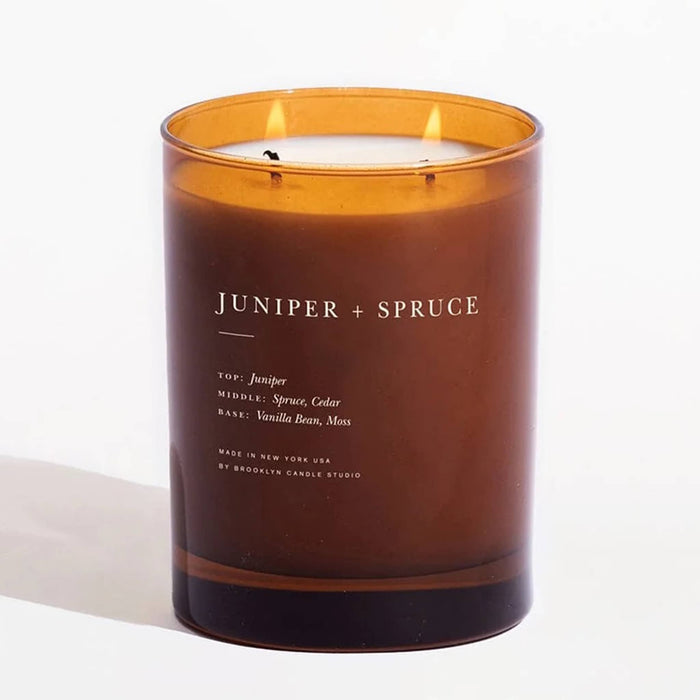 An amber glass jar candle with two lit wicks and white wax, labeled 'JUNIPER + SPRUCE' on a clean white background.