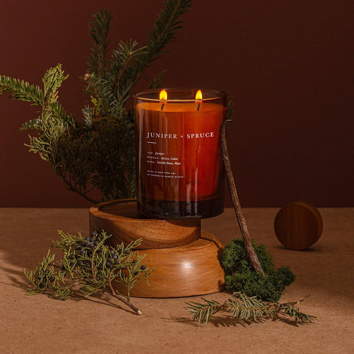 A lit double-wick candle in a dark amber glass jar sits on a wooden riser with evergreen branches, moss, and a small round wooden object around it against a warm brown background. The jar is labeled JUNIPER + SPRUCE.