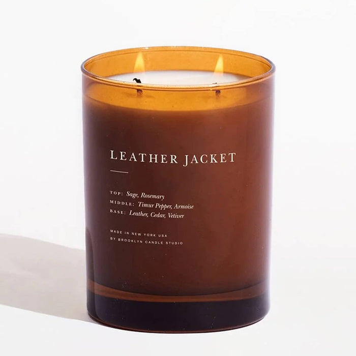 A lit amber glass jar candle with white wax and two visible flames. The front label reads 'LEATHER JACKET' with smaller scent notes below, photographed on a clean white background.