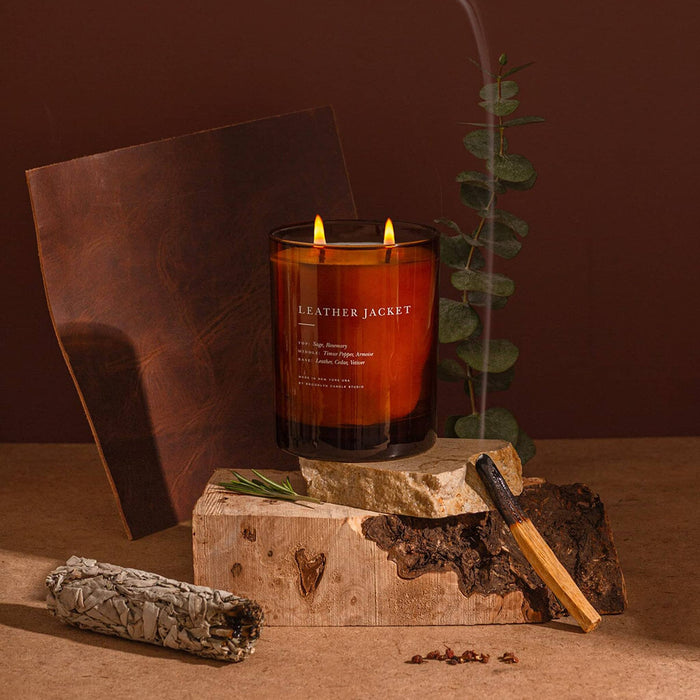 A lit Leather Jacket candle in a dark amber glass jar sits on a rustic wooden block, surrounded by dried botanicals, bark, a smudge bundle, and a leather sheet against a warm brown background.