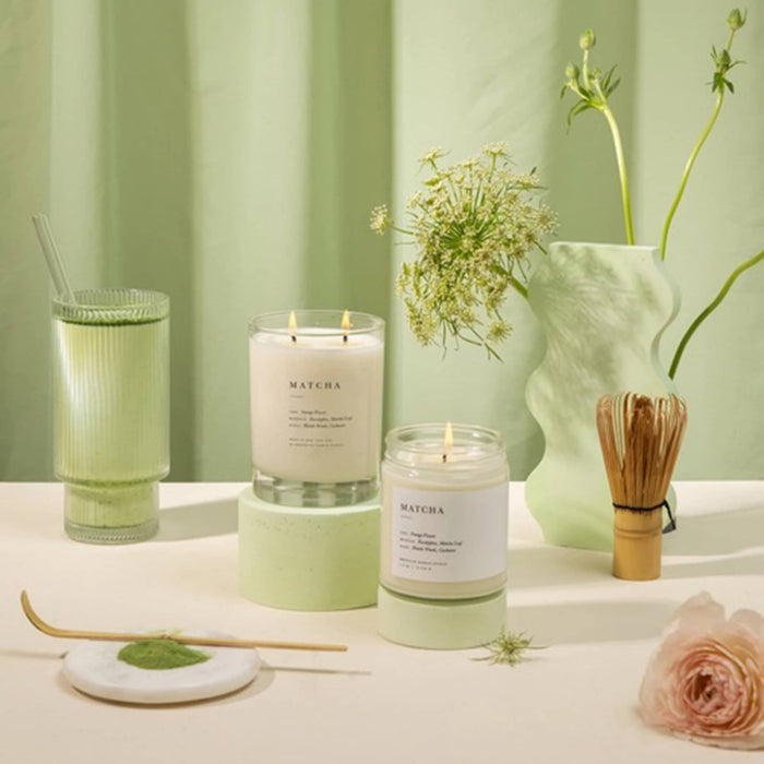 Two Matcha candles on a table with a green curtain and decorative items in the background.