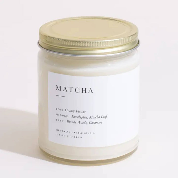 Brooklyn Candle Studio Matcha scented soy wax candle in a clear glass jar with a screw-on gold lid, on a white background.