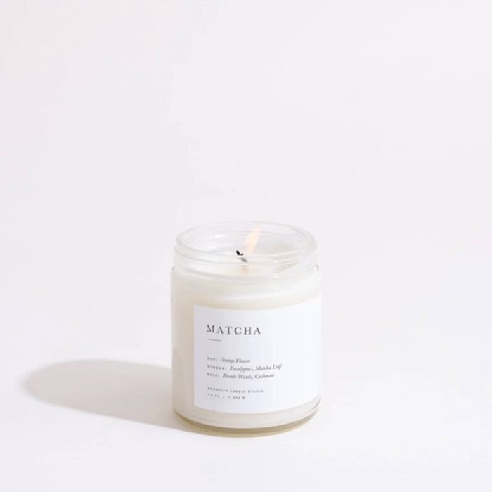 Brooklyn Candle Studio Matcha scented soy wax candle in a clear glass jar, wick lit, on a white background.