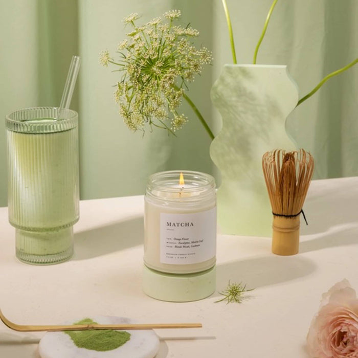 Matcha candle on a table with a glass of matcha, flowers, and a whisk.