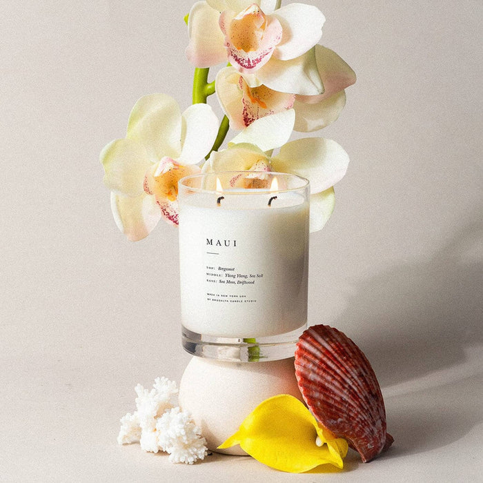 Candle with 'Maui' branding surrounded by flowers and a shell on a neutral background.