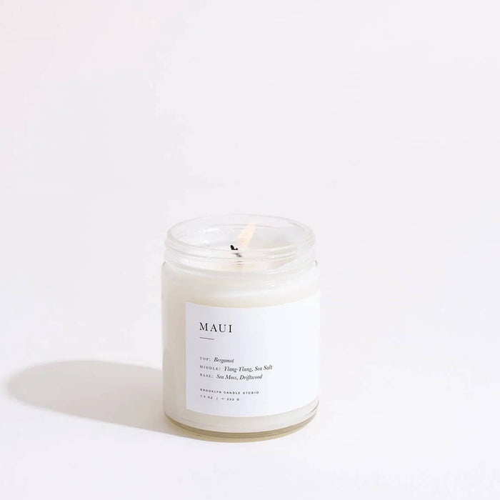 Candle in a jar labeled 'MAUI' with wick lit, on a white background.