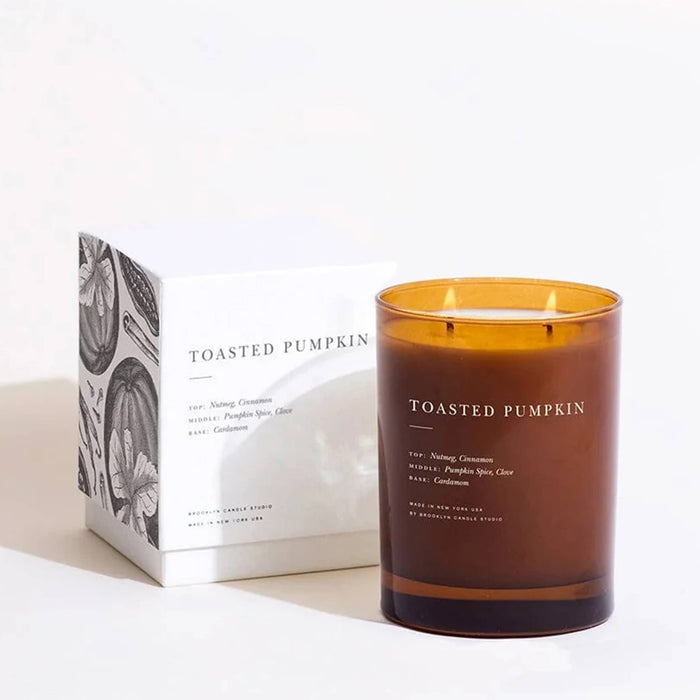 A brown amber glass jar candle labeled 'TOASTED PUMPKIN' stands next to its white box with black and white pumpkin illustrations on a clean white background.
